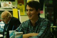 a man in a plaid shirt is sitting at a table with two cups of soda and a straw .