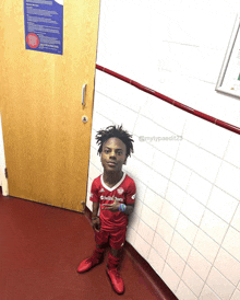 a young boy wearing a red adidas jersey stands in front of a white tiled wall