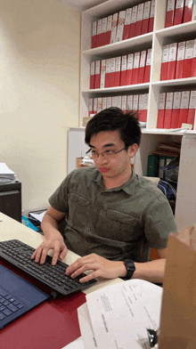 a man sits at a desk typing on a keyboard with a binder full of red binders on the shelf behind him