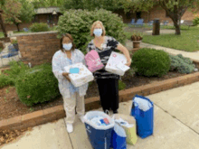 two women wearing masks are standing next to each other on a sidewalk holding gifts .