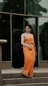 a woman wearing an orange saree and a white blouse is standing on stairs in front of a building .