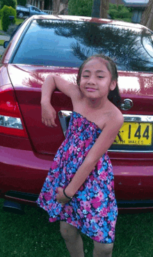 a girl in a floral dress is standing in front of a red car with a license plate that says 144