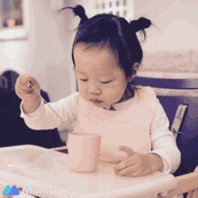 a little girl is sitting in a high chair with a cup and spoon in front of a momento photo