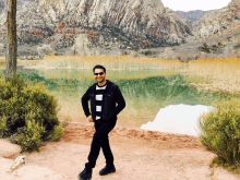 a man wearing sunglasses stands in front of a lake with mountains in the background