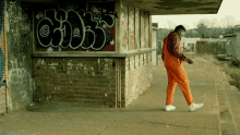 a man in orange overalls walks in front of a brick building with graffiti on it that says " chick "