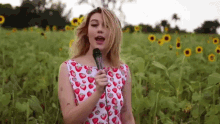 a woman in a dress with strawberries on it is singing into a microphone in a field of sunflowers .