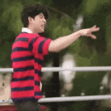 a young man in a red and blue striped shirt is standing in front of a fence and reaching out his hand .