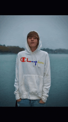 a man wearing a white champion hoodie is standing in front of a body of water .