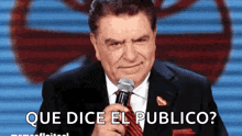 a man in a suit and tie is holding a microphone with the words que dice el publico below him