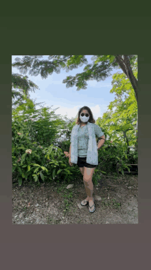 a woman wearing a mask and flip flops stands in front of a bush