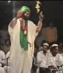 a man in a white robe and green turban is standing in front of a microphone and giving a thumbs up .