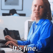 a woman in a blue shirt is sitting at a desk typing on a keyboard and says it 's fine .