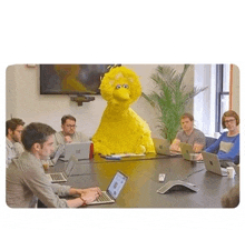 a group of people are sitting around a table with laptops and a giant sesame street character sitting on the table .