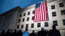 a group of people are standing in front of a large american flag hanging from the side of a building .