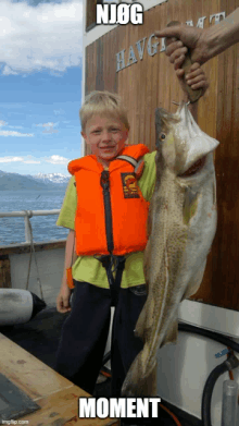 a young boy in an orange life vest holds a large fish in front of a building that says ' njog ' on it