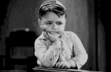 a black and white photo of a young boy sitting at a table with his hand on his chin .