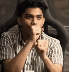 a young man in a striped shirt is sitting in a chair with his hands on his mouth .