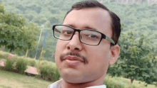 a man wearing glasses and a mustache looks at the camera with a mountain in the background