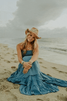 a woman wearing a blue dress and a straw hat sits on the beach