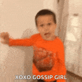 a young boy in an orange shirt is standing in front of a white wall and saying xoxo gossip girl .