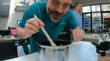 a man in a scrub top stirs a pot with a wooden spoon and the bbc logo is visible in the background