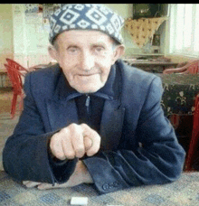 an elderly man wearing a hat and a suit is sitting at a table with a cigarette in his hand .