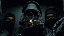 a man in a hood is holding a lighter in front of his face while standing next to two other men .