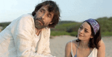 a man with a beard and a woman in a bikini are sitting on the beach looking at each other .