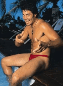 a shirtless man in red swim trunks is sitting on a ledge next to a pool