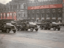 a row of military vehicles are driving down a street in front of a building with a banner that says " lenin "