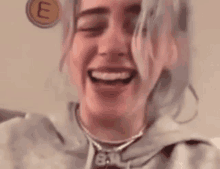 billie eilish is laughing with her eyes closed and wearing a necklace with the letter e on it .