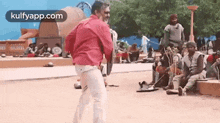 a man in a red shirt and white pants is standing in front of a group of people sitting on the ground .