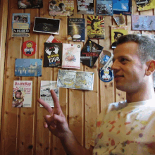 a man giving a peace sign in front of a wall covered in stickers including one that says boom bap