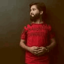 a man with a beard wearing a red t-shirt is standing in front of a wall .