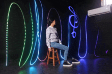 a woman sits on a stool in front of a wall with glowing lights behind her