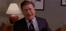 a man in a suit and tie is sitting on a couch with his eyes closed and laughing .