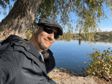 a man wearing sunglasses and a hat sits under a tree near a lake