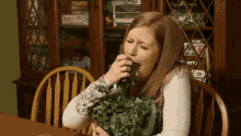 a woman is sitting at a table with a bowl of greens in front of a bookcase with twenty-one on it