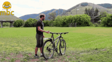 a man stands next to a bicycle in a grassy field with mountains in the background and the word mbc in the corner