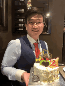 a man in a vest and tie holds a cake that has the number 2 on it