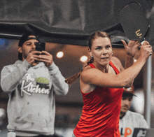 a woman in a red tank top is swinging a paddle while a man takes a picture of her