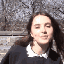 a young woman wearing a blue sweater and a white collared shirt is standing in front of a stone wall .