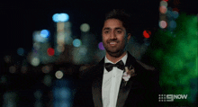 a man in a tuxedo and bow tie is standing in front of a city skyline at night ..