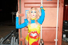 a drag queen in a yellow and blue outfit is standing in front of a red container with a blue sticker on it