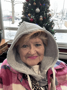 an elderly woman wearing a hoodie stands in front of a christmas tree and a window