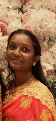 a woman in a red and gold sari is standing in front of a wall of flowers and smiling .