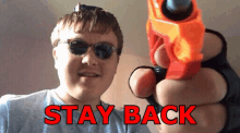 a man wearing sunglasses and gloves is holding a toy gun and says " stay back " in red