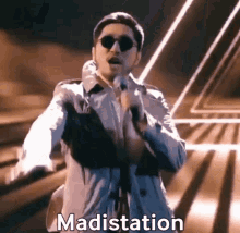 a man wearing sunglasses and a trench coat is singing into a microphone with the word maditation written below him .