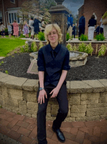 a young man in a black vest and tie is sitting on a stone wall in front of a fountain