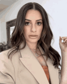 a woman wearing a tan blazer and a necklace looks at the camera